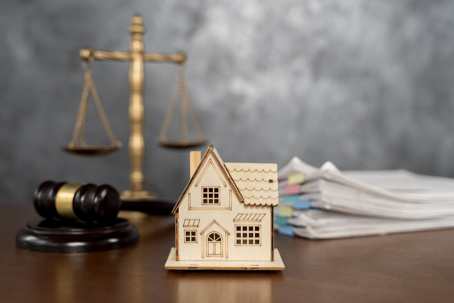 Wooden house model placed before a gavel, justice scale, and document stack, illustrating property law, mortgage issues, legal disputes, ownership rights, and real estate paperwork.